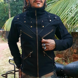 May include: A black long-sleeve jacket featuring a high collar, a central brass-toned zip, and multiple diagonal zipped pockets. The shoulders are adorned with vertical fabric strips accented by small metallic studs and hexagonal rivets.