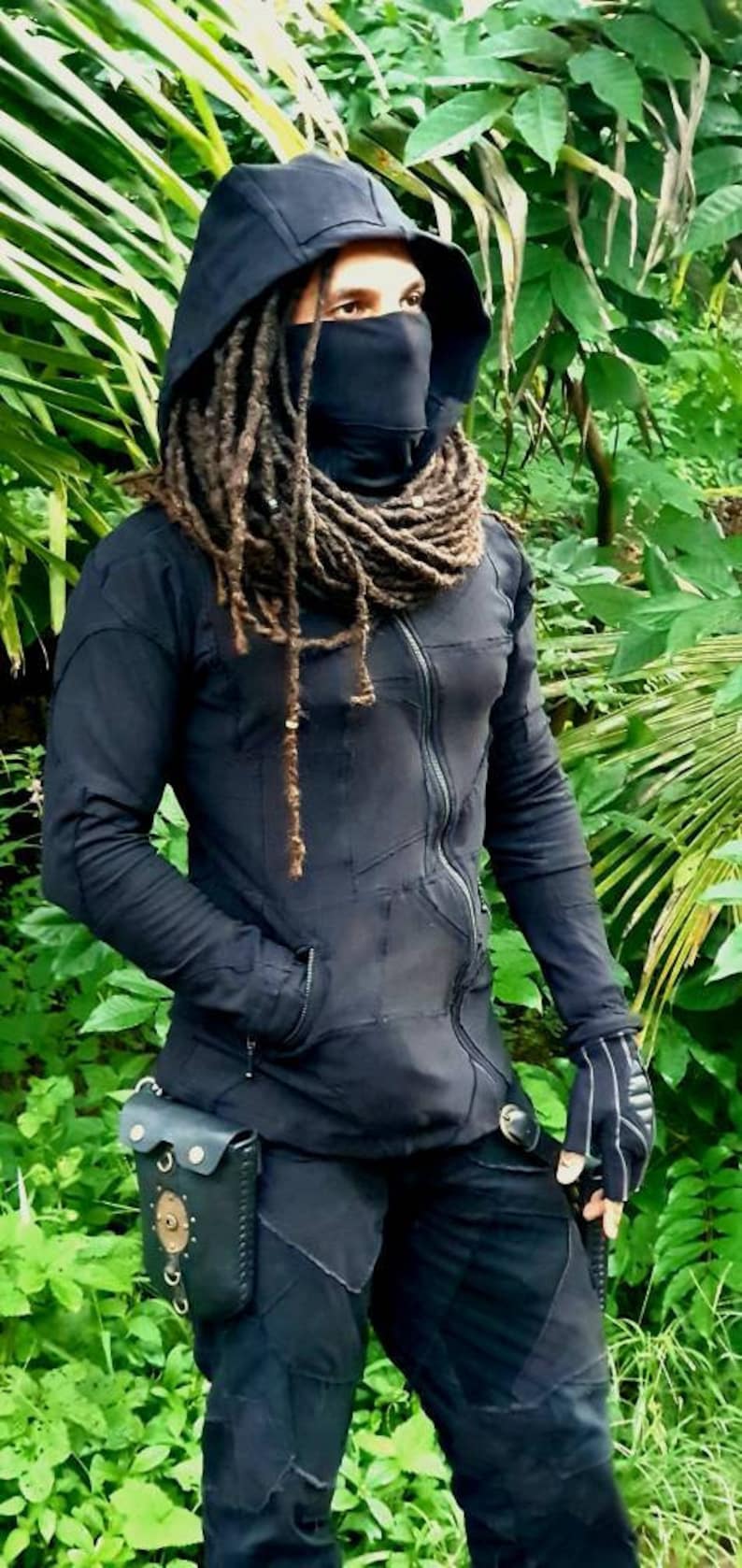 May include: A person wearing a black hooded jacket with a black face mask and long dreadlocks. The jacket has a zip and pockets. The person is wearing black trousers and a leather belt with a pouch.
