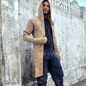 May include: A long, tan hooded coat with a repeating circular pattern. The coat is worn over a black shirt and dark blue, textured pants. The background is a weathered white wall and a blue sky.