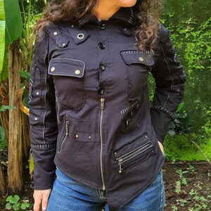 Puede incluir: Una chaqueta negra de manga larga con cierre de cremallera frontal, m&uacute;ltiples bolsillos con botones y detalles de costuras decorativas. La prenda incluye ojales met&aacute;licos, herrajes plateados y un cuello estructurado, ofreciendo un aspecto utilitario con costuras visibles y paneles de tela superpuestos.