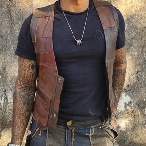 May include: A sleeveless vest constructed from multiple panels of brown leather in varying shades, featuring a front button closure and visible stitching. The vest is worn over a plain black t-shirt, paired with dark blue trousers that have a central zipper.