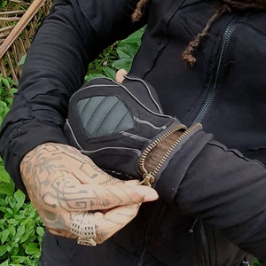 May include: A black jacket with a zip closure and multiple pockets. The jacket has a distressed look with visible stitching and seams. The zip is partially open, revealing a black and grey inner lining.