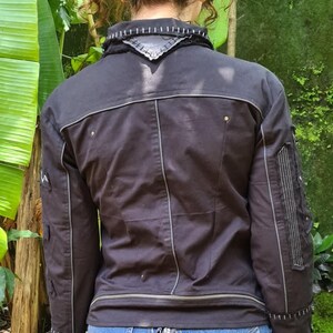 Puede incluir: Una chaqueta negra con costuras blancas en contraste, cuello alto con un parche triangular y mangas largas con paneles verticales cosidos. La chaqueta incluye un detalle de cremallera horizontal en la cintura y herrajes met&aacute;licos plateados.