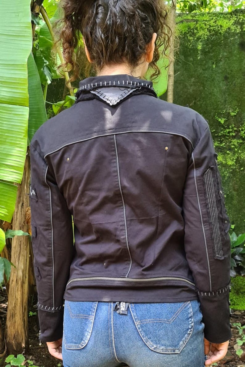 Puede incluir: Una chaqueta negra con costuras blancas en contraste, cuello alto con un parche triangular y mangas largas con paneles verticales cosidos. La chaqueta incluye un detalle de cremallera horizontal en la cintura y herrajes met&aacute;licos plateados.
