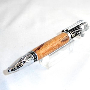 Op de afbeelding: Een handgemaakte pen met een lichtbruine houten schacht en zilverkleurige metalen accenten. De pen heeft een uniek ontwerp met een veermechanisme aan de bovenkant en een facetgeslepen punt. De pen wordt gepresenteerd op een transparante acryl standaard.