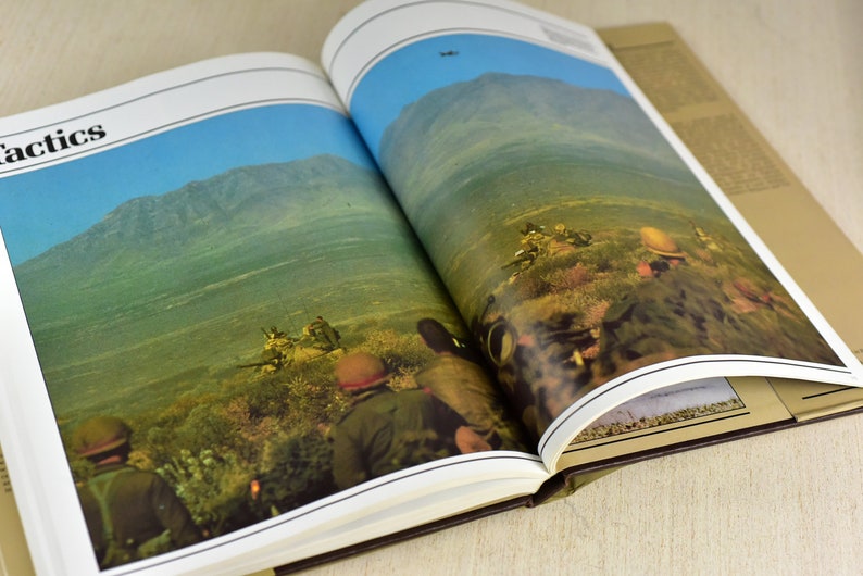 May include: An open book with a photograph of soldiers in a military setting. The book's title is "Tactics".
