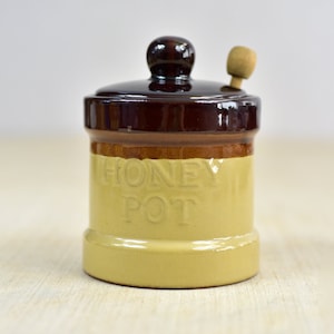 May include: A ceramic honey pot with a dark brown lid and base, and a tan body. The words "HONEY POT" are embossed on the front. A small wooden honey dipper rests on the lid. The pot is designed for storing honey.