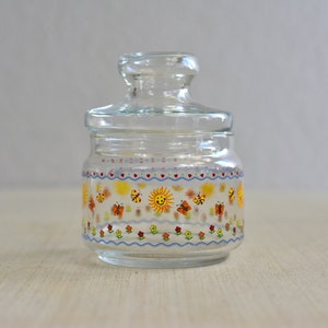 Puede incluir: Un tarro de vidrio transparente con tapa, decorado con un patrón colorido de mariposas, flores y un sol sonriente. El tarro tiene un borde azul, blanco y amarillo.