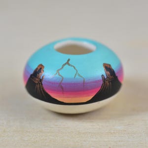 May include: A small, round ceramic vase with a turquoise top and a white base. The vase features a painted landscape with black figures and a colourful sunset design in shades of pink, orange, and purple.
