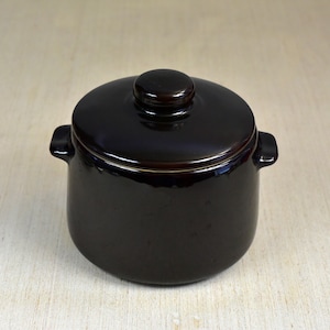 May include: A small, dark brown ceramic pot with a lid and two handles. The pot has a light brown rim around the top.