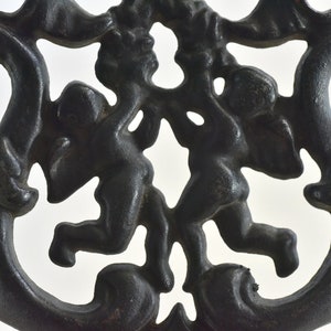 Puede incluir: Diseño decorativo de metal negro con dos querubines con detalles intrincados. Los querubines están colocados de espaldas, creando un diseño simétrico.