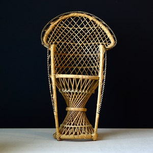 May include: A small, light brown wicker chair featuring a high, rounded back with a diamond-patterned lattice weave. The seat is circular, supported by a flared, hourglass-shaped base constructed from vertical and diagonal woven reed strands.