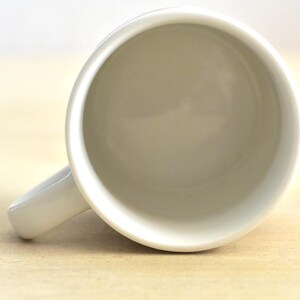 K&ouml;nnte beinhalten: Eine wei&szlig;e Keramik-Kaffeetasse mit gebogenem Henkel. Die Tasse ist leer und von oben betrachtet, wodurch ein glattes, sauberes Inneres sichtbar wird. Die Tasse steht auf einer hellen Holzoberfl&auml;che.