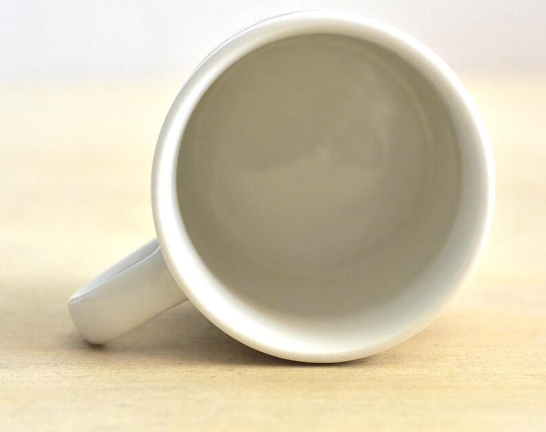 K&ouml;nnte beinhalten: Eine wei&szlig;e Keramik-Kaffeetasse mit gebogenem Henkel. Die Tasse ist leer und von oben betrachtet, wodurch ein glattes, sauberes Inneres sichtbar wird. Die Tasse steht auf einer hellen Holzoberfl&auml;che.