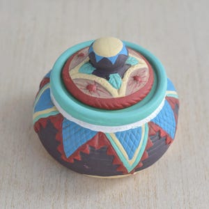 May include: A small, round, ceramic jar with a lid. The jar is decorated with a colourful geometric pattern in shades of blue, red, yellow, and brown. The lid has a similar pattern and a small, round knob on top.