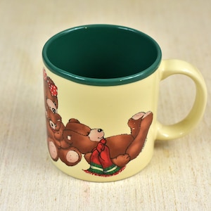 Op de afbeelding: Een keramische mok met een groene rand en een crèmekleurig lichaam. De mok heeft een cartoonillustratie van twee bruine teddyberen die rode en groene kerstkledij dragen.