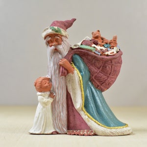 May include: A ceramic Santa Claus figurine with a long white beard, red hat, and a sack full of toys. A small child in a white robe stands beside Santa. The figurine has a teal and gold trim on the robe.