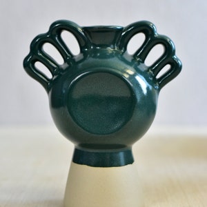 Pode incluir: Um vaso de cer&acirc;mica verde com base branca e quatro al&ccedil;as. O vaso tem um corpo redondo e uma abertura larga.
