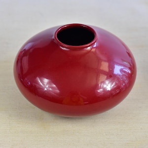 Peut inclure: Un vase en céramique rouge brillant de forme ovale arrondie. Le vase a un corps large et une petite ouverture en haut, avec un intérieur sombre. La surface réfléchit la lumière, créant des reflets. Ce vase décoratif convient à la décoration intérieure.