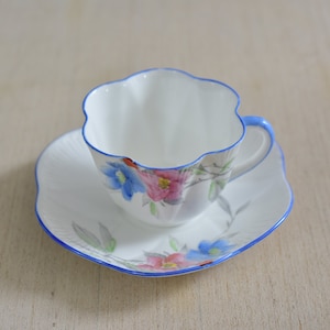 May include: A white teacup and saucer with a blue rim and floral pattern. The cup has a scalloped edge and the saucer has a scalloped rim.