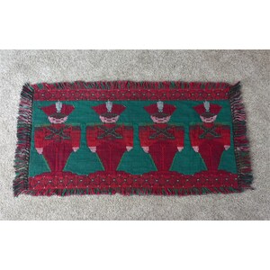 Puede incluir: Textil rectangular tejido con un patrón repetido de cuatro soldados de juguete con uniformes y sombreros rojos sobre un fondo verde. El textil tiene un borde y flecos rojos. Los soldados tienen caras blancas y sostienen rifles.