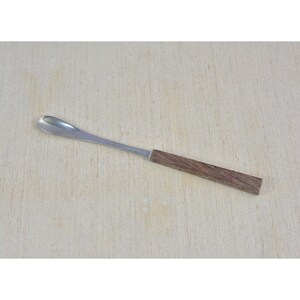Puede incluir: Una cuchara de acero inoxidable con mango de madera. La cuchara tiene un extremo redondeado y está diseñada para recoger.