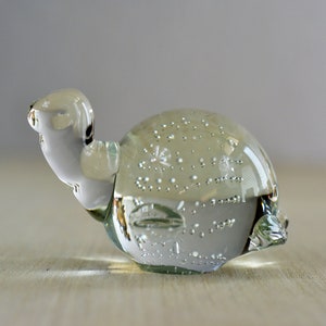 Puede incluir: Una figurita de tortuga de vidrio transparente con burbujas de aire atrapadas en su interior. La tortuga está sentada sobre una superficie de madera clara.
