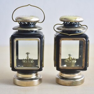 May include: Two antique black and brass lanterns with clear glass panels. Each lantern has a handle, a decorative top, and a base. The lanterns have a vintage aesthetic and are likely used for decorative purposes.