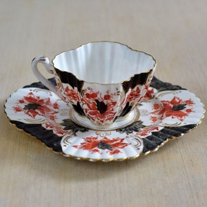 Puede incluir: Una taza de té y un platillo blancos con un patrón floral negro y rojo. La taza tiene un borde festoneado y el platillo tiene un borde festoneado.