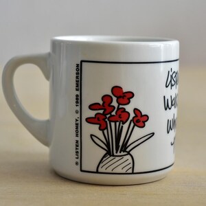 K&ouml;nnte beinhalten: Wei&szlig;er Keramikbecher mit schwarzem Henkel. Der Becher zeigt eine Cartoon-Illustration von roten Blumen in einer Vase, mit dem Text "listen, watch, wait" und den Urheberrechtsinformationen.