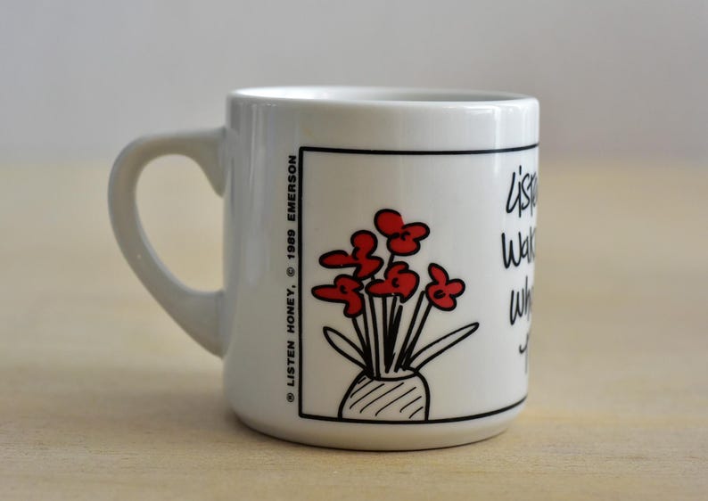 K&ouml;nnte beinhalten: Wei&szlig;er Keramikbecher mit schwarzem Henkel. Der Becher zeigt eine Cartoon-Illustration von roten Blumen in einer Vase, mit dem Text "listen, watch, wait" und den Urheberrechtsinformationen.