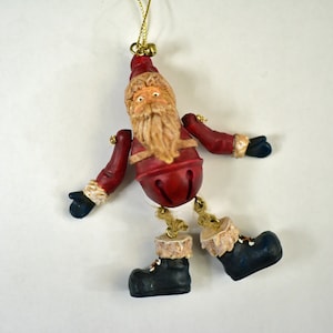 May include: A Christmas ornament shaped like Santa Claus. The ornament has a red body and arms, a white beard, and black boots. The arms and legs are jointed, and the ornament is suspended by a gold string.