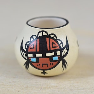 May include: A small, round, cream-colored ceramic pot with a hand-painted Native American-inspired design. The design features a stylized face with black, red, and turquoise accents. The pot has a wide opening and a smooth, glossy finish.