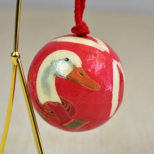 Puede incluir: Adorno navideño redondo rojo con una ilustración de pato blanco y rayas blancas. El pato tiene un pico naranja y lleva una bufanda roja. El adorno cuelga de una cuerda roja.