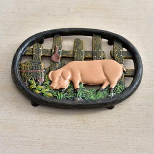 May include: An oval cast iron trivet featuring a raised relief design of a pink pig standing in green grass. Behind the pig is a brown wooden fence and a barrel. The entire scene is framed by a thick, dark, textured black metal border.