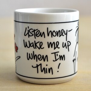 K&ouml;nnte beinhalten: Wei&szlig;e Keramiktasse mit schwarzem Text: &bdquo;Listen honey - Wake me up when I'm thin!&ldquo; Die Tasse hat einen schwarzen Rand und Boden, mit einem einfachen Blumen- und Strichdesign. Die Tasse ist zylindrisch.