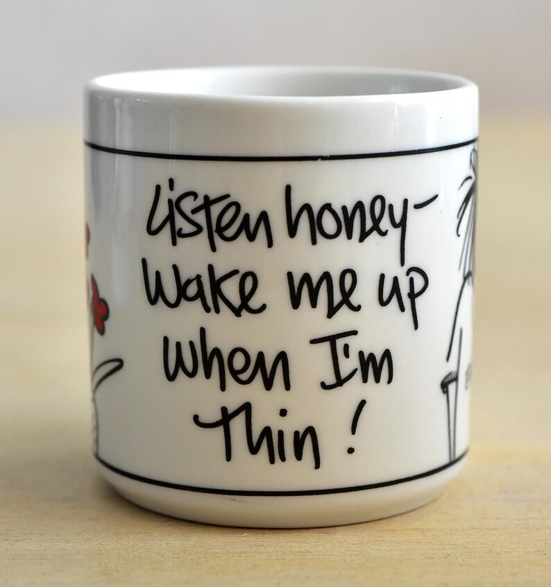 K&ouml;nnte beinhalten: Wei&szlig;e Keramiktasse mit schwarzem Text: &bdquo;Listen honey - Wake me up when I'm thin!&ldquo; Die Tasse hat einen schwarzen Rand und Boden, mit einem einfachen Blumen- und Strichdesign. Die Tasse ist zylindrisch.