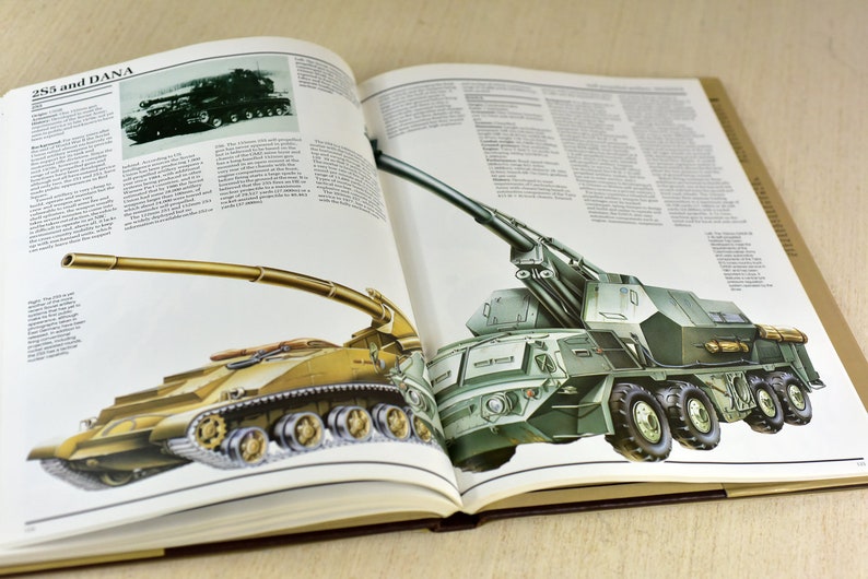 May include: An open book with illustrations of two military vehicles. The book is titled "255 and DANA". The first vehicle is a green self-propelled howitzer with a long barrel and six wheels. The second vehicle is a tan self-propelled howitzer with a long barrel and tracks.
