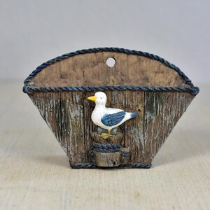 Puede incluir: Un bolsillo de pared decorativo de madera con temática náutica. El bolsillo tiene forma de semicírculo y presenta una figura de gaviota. La gaviota es blanca con alas azules y pico amarillo. El bolsillo es marrón con ribete de cuerda.