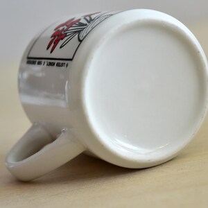 K&ouml;nnte beinhalten: Wei&szlig;e Keramiktasse mit gebogenem Henkel, mit einem floralen Design in Rot und Schwarz. Der Text "LISTEN HONEY, &copy; 1990" ist auf der Seite aufgedruckt. Die Tasse liegt auf einer hellen Holzoberfl&auml;che.