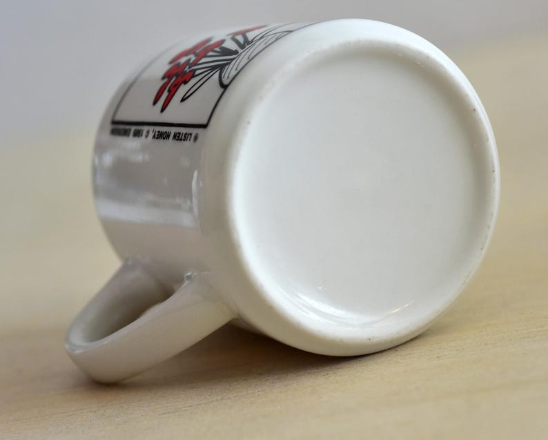 K&ouml;nnte beinhalten: Wei&szlig;e Keramiktasse mit gebogenem Henkel, mit einem floralen Design in Rot und Schwarz. Der Text "LISTEN HONEY, &copy; 1990" ist auf der Seite aufgedruckt. Die Tasse liegt auf einer hellen Holzoberfl&auml;che.