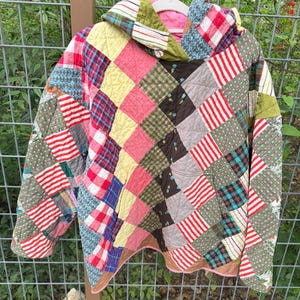 May include: A patchwork hoodie with a variety of colorful fabric squares. The jacket features a mix of patterns, including stripes, plaid, and floral designs. The hood is lined with pink fabric, and the jacket is hanging on a white hanger.