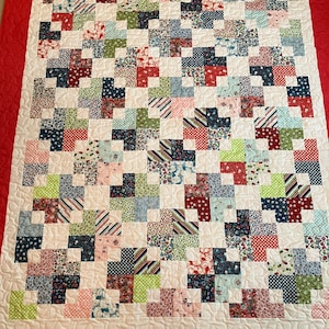 May include: A white quilt with a red border. The quilt is made up of many small squares of different fabrics, including floral prints, stripes, and polka dots. The squares are arranged in a checkerboard pattern.