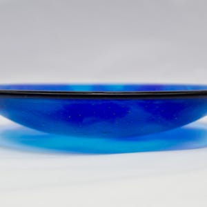 May include: A striking blue glass bowl with a black rim. The bowl has a shallow, rounded form and is set against a white backdrop. The colour transitions from a deep blue to a lighter shade, creating a gradient effect.