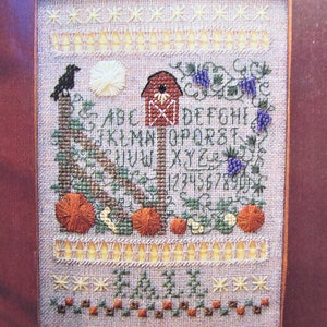 Puede incluir: Muestra de punto de cruz con tema otoñal. Presenta un cuervo, calabazas, una casa para pájaros y vides. Incluye el alfabeto y números. La muestra está enmarcada en un borde marrón.