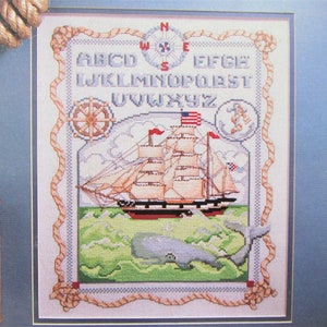 Puede incluir: Un diseño de punto de cruz que presenta un barco con una bandera estadounidense navegando en el océano con una ballena nadando debajo. El alfabeto está bordado sobre el barco con una brújula y un ancla.
