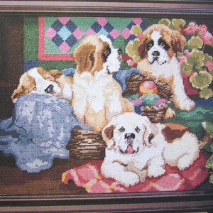 Op de afbeelding: Een ingelijst borduurwerk met kruissteek, met vier schattige Sint-Bernarduspups. De pups zijn afgebeeld in een knusse setting met een mand vol kleurrijke paaseieren en een roze-groen quilt.