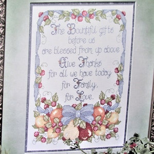 Puede incluir: Un bordado a punto de cruz enmarcado con fondo blanco y borde azul y verde. El bordado presenta un diseño floral con fruta y un lazo de cinta. El texto dice: "The Bountiful gifts before us are blessed from up above Give Thanks for all we have today for Family, for Love."