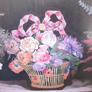 May include: A needlepoint wall hanging depicting a basket overflowing with colorful flowers, including roses, peonies, and daisies. A large pink and purple bow adorns the top of the basket.