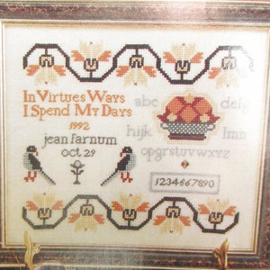 May include: A framed cross-stitch sampler with a white background and brown frame. The sampler features a floral border, two birds, a small plant, and the text "In Virtues Ways I Spend My Days 1992 jean farnum oct 29 abc del hijk Imn opgrstuvwxyz 1234667890".